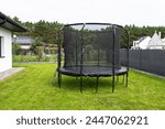 Large childrens jumping trampoline with protective net and closed zipper, standing in the garden, visible mesh masking the fence.