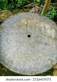 A large, circular, grey concrete object is centered in the frame, likely a stepping stone or a small garden table top. The surface is heavily textured with a distinct, dry-cracked pattern. A small, dark hole is present in the exact center. The object is situated outdoors, surrounded by green foliage, plants, and earth, suggesting a garden or natural setting.