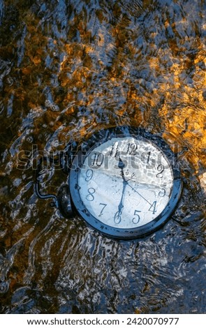 Large clock in a flowing stream with half of the clock face submerged. Concept image losing track out of time, relativity science, climate change, flowing time, with ripples distorting the numbers. 