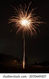 Large, colourful, bright firework display with orange rocket against a black background with crowd visible in the distance in a park on bonfire night