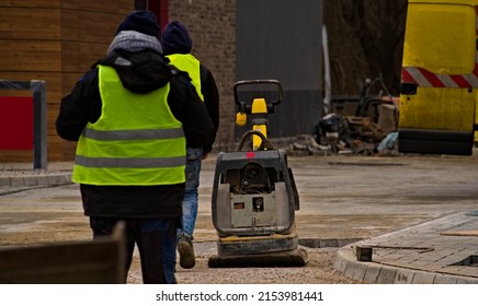 Large Compactor (over Two Hundred Kilos). Paving Large Areas Of The Future Car Park. After A Lunch Break, Employees Return To Work (wearing Reflective Protective Suits).