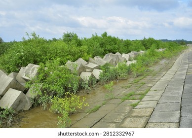 A large concrete embankment built with rectangular slabs forming a sloped spillway leading toward a water body. Vegetation grows near the water’s edge, and a small observation tower stands in the background under a partly cloudy sky. The scene highlights engineered flood‑control infrastructure integrated into a natural river landscape.