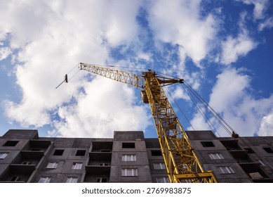 A large construction crane is positioned next to a partially built apartment building under a dynamic sky filled with clouds