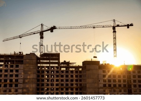 Large construction cranes tower over a buildings' framework as the sun sets in the background, casting warm hues and highlighting the ongoing development in a bustling city.