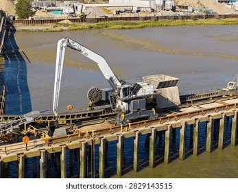 Large construction machine with a long arm moves sand on a wooden dock over a river during the bright daylight hours with industrial facilities in the background