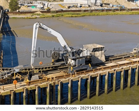 Large construction machine with a long arm moves sand on a wooden dock over a river during the bright daylight hours with industrial facilities in the background