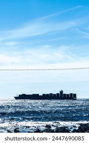 A large container ship is traversing the sunlit ocean waters, with the horizon visible in the distance. Whitecaps decorate the surface of the sea, indicating a brisk breeze