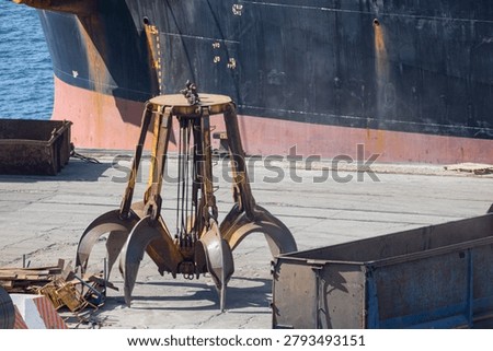 A large crane with claw-like attachments is positioned at a loading dock. It is ready to lift cargo from a nearby ship to the shore. The water reflects sunlight