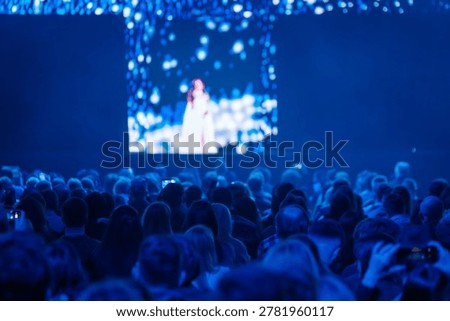 Large crowd watching live performance on stage and big screen, atmospheric blue lighting, audience using smartphones to record show.
