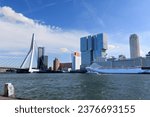 A large curise ship docked in Rotterdam in front of highrises on a sunny day. The Erasmus bridge is in the background.