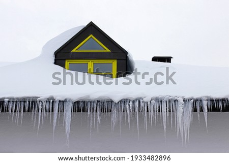 Large dangerous icicles and a roof avalanche on a house roof with yellow windows in winter 