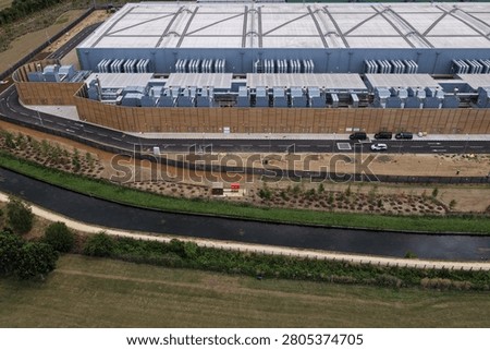 Large Data Center building stands by a peaceful canal in the countryside. The modern facility has visible cooling equipment. A road with parked vehicles runs past the building during daytime.