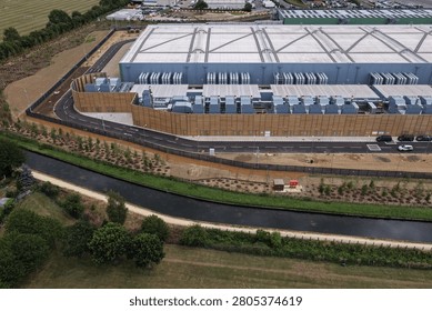 Large data center structure seen from above, featuring extensive cooling systems along its side. A narrow canal and lush fields run alongside the secure facility under a bright sky.