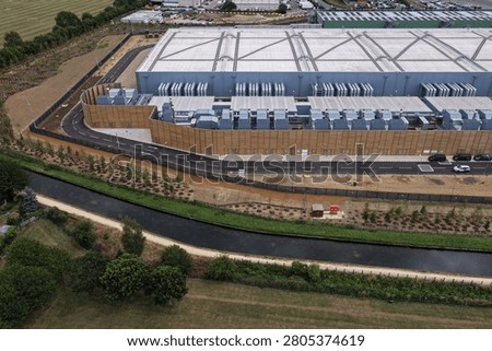 Large data center structure seen from above, featuring extensive cooling systems along its side. A narrow canal and lush fields run alongside the secure facility under a bright sky.
