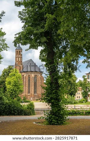 A large deciduous tree frames the view of a historic brick church with a clock tower, set in a green park under a changeable sky
