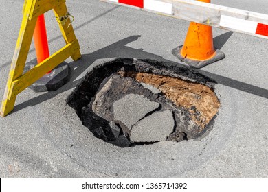 A large, deep pothole in a road. Part of a traffic cone and sawhorse visible blocking the hole.