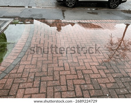Large deep puddle after heavy rain on a driveway. The water reflects a house opposite and a tree on the pavement. 
