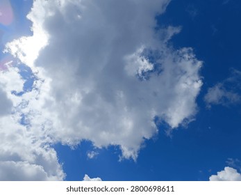 A large, dense white and grey cloud dominates the center of the frame, partially obscuring the sun. Bright sunlight and colorful lens flares peek out from behind the upper left edge of the cloud. The surrounding sky is a vibrant blue.