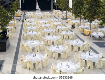 Large Dining Table Set For Wedding, Dinner Or Another Corporate Event With Beautiful Decoration Between Green Trees Inside With People Moving Blurred