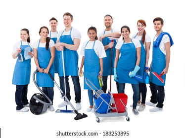 Large diverse group of janitors wearing blue aprons standing grouped together with their equipment smiling at the camera  isolated on white
