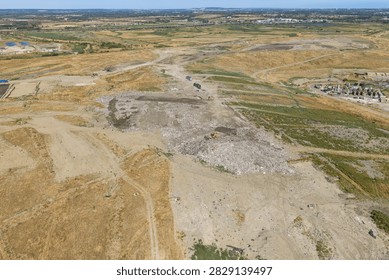 Large dump site with heavy construction vehicles pushing and organizing waste materials under the clear daylight sky in an open industrial area surrounded by dry grassy fields and roads