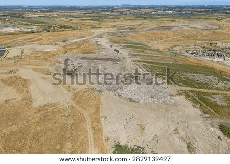 Large dump site with heavy construction vehicles pushing and organizing waste materials under the clear daylight sky in an open industrial area surrounded by dry grassy fields and roads
