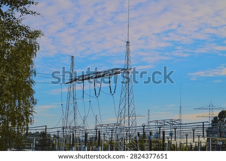 A large electrical substation featuring multiple high-voltage transmission towers and power lines is photographed under a vivid blue sky with scattered white clouds. Green trees are visible on the left edge of the frame. Ideal for concepts related to energy infrastructure, power generation, and industrial utility grids.