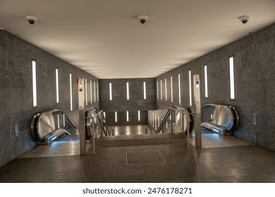 A large, empty, modern-looking subway station with escalators and stairs. The escalators are silver and the stairs are also silver