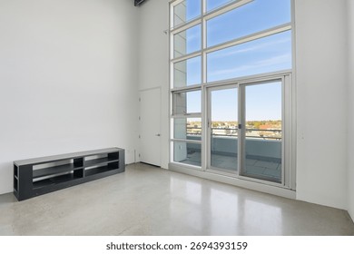 Large, empty room with a window overlooking a city. The room is clean and uncluttered, with a black couch and a black shelf. The window lets in natural light and offers a view of the city below