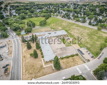 Large empty lot with a school building in the middle. The lot is surrounded by a fence and has a parking lot with several cars
