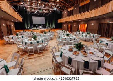 Large event hall with elegantly set round tables, floral centerpieces, food, and a stage prepared for a performance or formal gathering.
