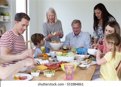 Large Family Are Sat Round A Table In The Dining Room Of A Home. They Are Eating Mediterranean Style Food.