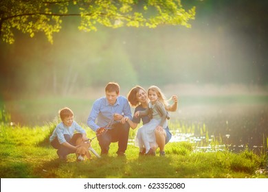 A Large Family Is Walking His Beagle In A Park By The Lake. Mom Enjoys The Hair Of Her Daughter, And Her Father And Son Train The Dog