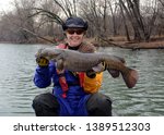 A large fat brown flathead catfish being held horizontally by a smiling woman sitting in a canoe in a blue and gold dry suit on a blue river in winter.