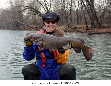 A large fat brown flathead catfish being held horizontally by a smiling woman sitting in a canoe in a blue and gold dry suit on a blue river in winter.
