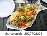 Large fish platter with various fried fish fillets, vegetables, lemon, herbs and sauce, served with white plates on a dark wooden table, seen from above, selected focus