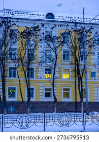 Large flock of crows gathering on leafless trees and a snow-covered building roof in a cold urban winter setting, with illuminated windows adding a warm glow from inside