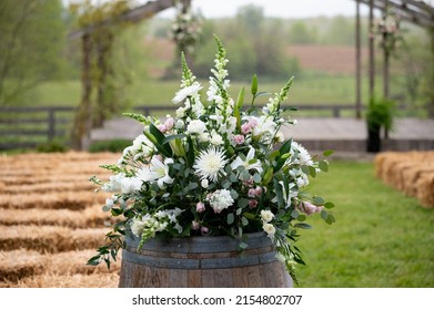 Large Floral Arrangement Outdoors For Wedding Ceremony Sight. White And Green Colors Decorate The Wood Barrel Top. Outside Event Centerpiece. 