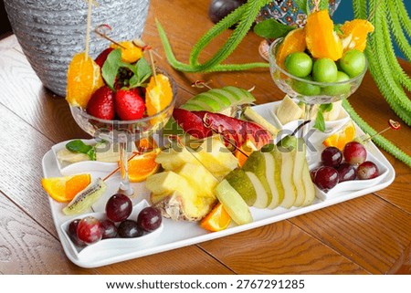 A large fruit platter is set on a wooden table. It includes strawberries, grapes, oranges, apples, and pears. The fruits are arranged in bowls and on plates.