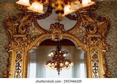 A Large Gold Chandelier With Patterns And Many Shades Is Reflected In A Large Antique Wall Mirror With A Gilded Carved Frame.