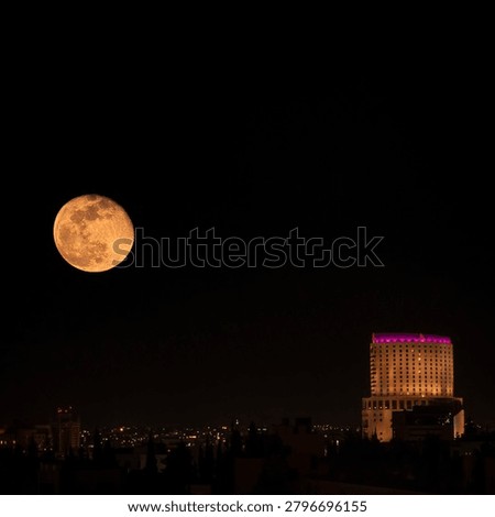 A large golden moon hangs dramatically over the dark skyline of Amman, creating a striking contrast between the natural glow of the night sky and the illuminated city below. The building on the right, lit with warm and purple tones, anchors the composition and gives the image a modern urban character. The deep black sky allows the moon to become the emotional center of the photograph, while the scattered city lights in the distance add depth and atmosphere. The scene feels calm, cinematic, and slightly mysterious, capturing Amman at a quiet and magical moment after sunset.