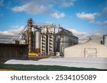 large grain elevator with multiple cylindrical metal silos, structural elements, and a complex system of pipes. It stands against a blue sky, with a field in the foreground after harvest