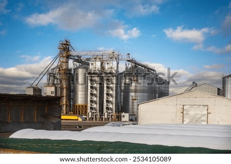 large grain elevator with multiple cylindrical metal silos, structural elements, and a complex system of pipes. It stands against a blue sky, with a field in the foreground after harvest