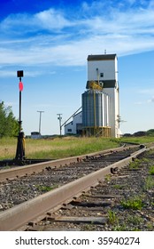 A large grain elevator in the prairies is situated near some empty railroad tracks, shot with a nice blue sky