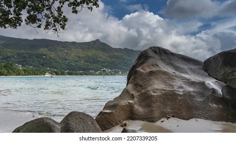 Large Granite Boulders On A Tropical Beach. The Turquoise Ocean Is Calm. A Green Hill In The Distance. Tree Branches Against The Sky And Clouds. Seychelles. Mahe