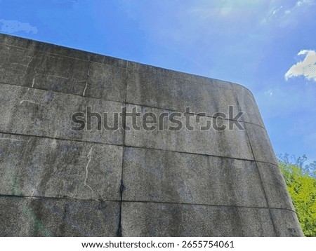 Large gray concrete wall structure stands against a bright blue sky backdrop with some foliage, creating a weathered yet modern architectural scene suitable for urban design or construction visuals.