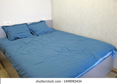 A Large Gray Double Bed Is In The Corner Of A Light Gray Room. The Bed Is Covered With Monochromatic Blue Linen, Two Pillows On The Bed And Two Sockets On The Wall. Crumpled Empty Bed In The Morning.