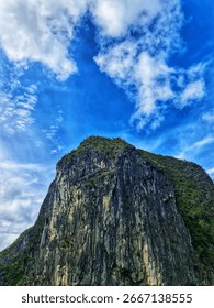 A large, gray mountain rises dramatically against a bright blue sky filled with puffy white clouds, creating a stunning landscape perfect for travel and outdoor adventure marketing.