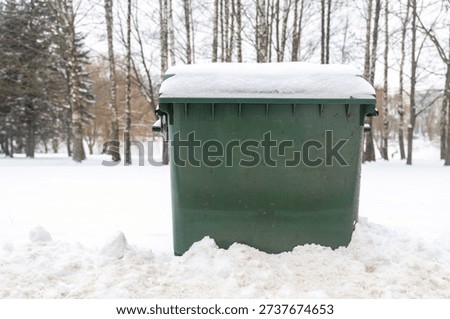 A large green garbage container against the background of trees in a snow-covered city park.