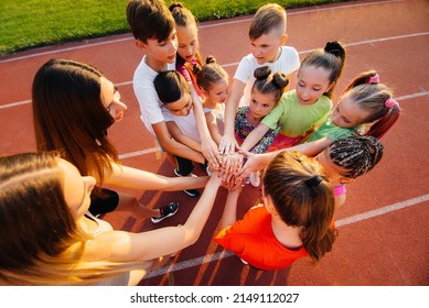 A Large Group Of Children, Boys And Girls, Stand Together In A Circle And Fold Their Hands, Tuning Up And Raising Team Spirit Before The Game At The Stadium During Sunset. A Healthy Lifestyle.